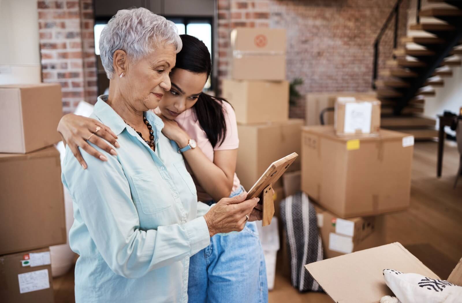 A young adult helping an older adult pack their home, taking a moment to reminisce over a photograph and hugging.