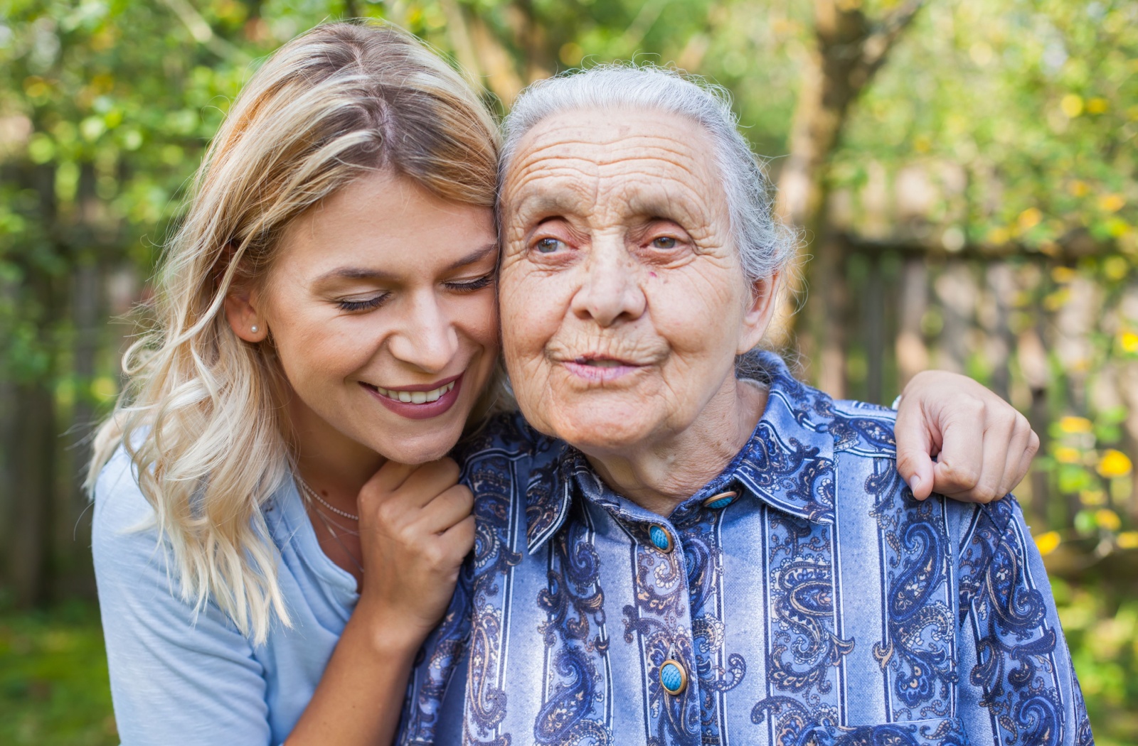 An adult grandchild hugs their older grandparent around the shoulders during a visit to their memory care home