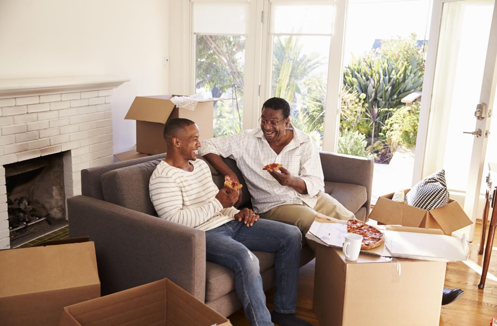 An adult child takes a pizza break with their older parent while packing to prepare them to move into memory care
