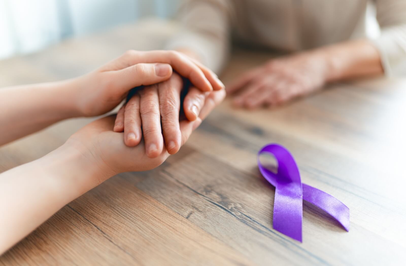 Someone holds a senior’s hand on top of a wooden table next to a purple ribbon representing Alzheimer’s awareness