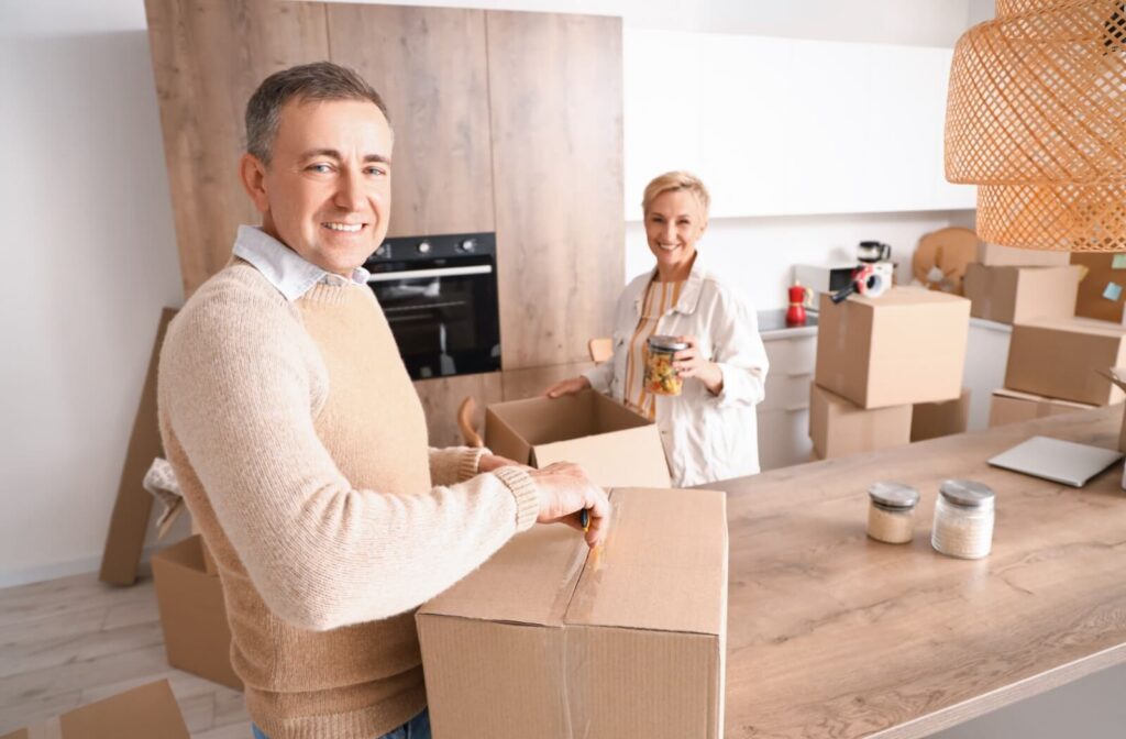 A senior couple in beige smile as they pack up unneeded items from their kitchen to donate prior to a move to assisted living