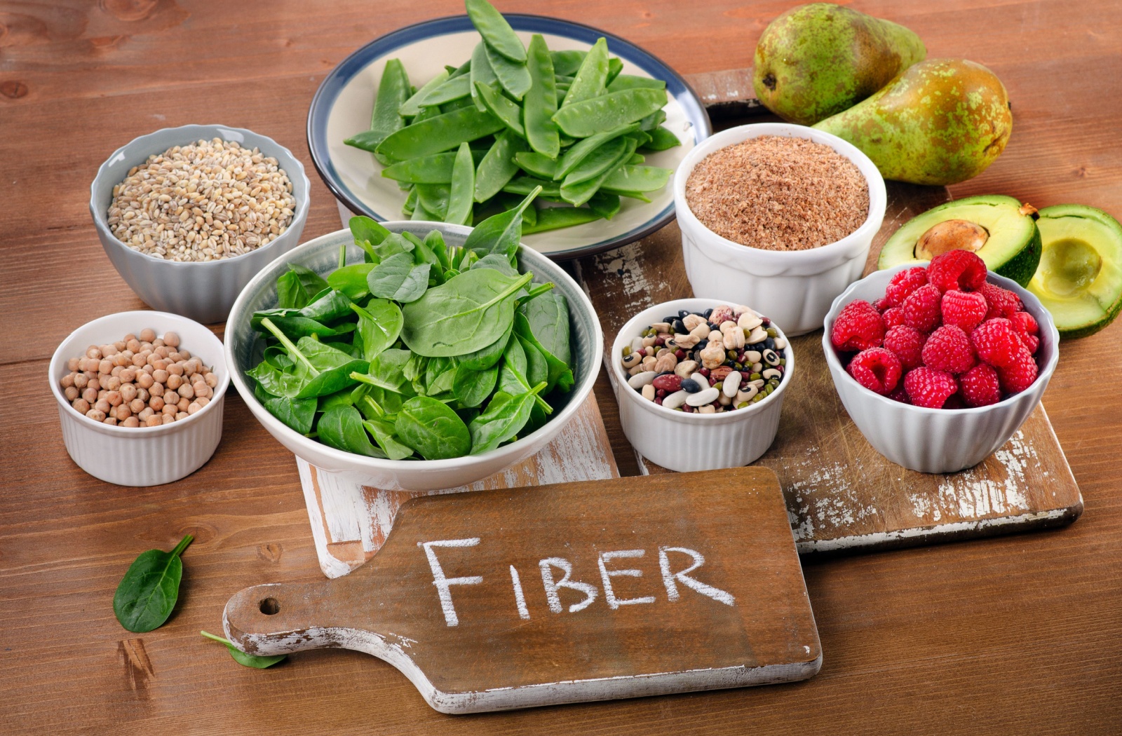 A collection of bowls of fiber-heavy foods, including greens, nuts, and raspberries, sit on a table with a “fiber” label