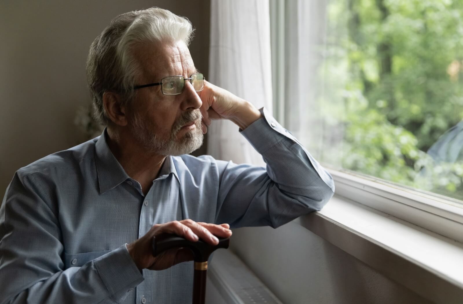 A senior with alzheimer's sitting with a cane in hand stares out the window on an overcast day looking distressed