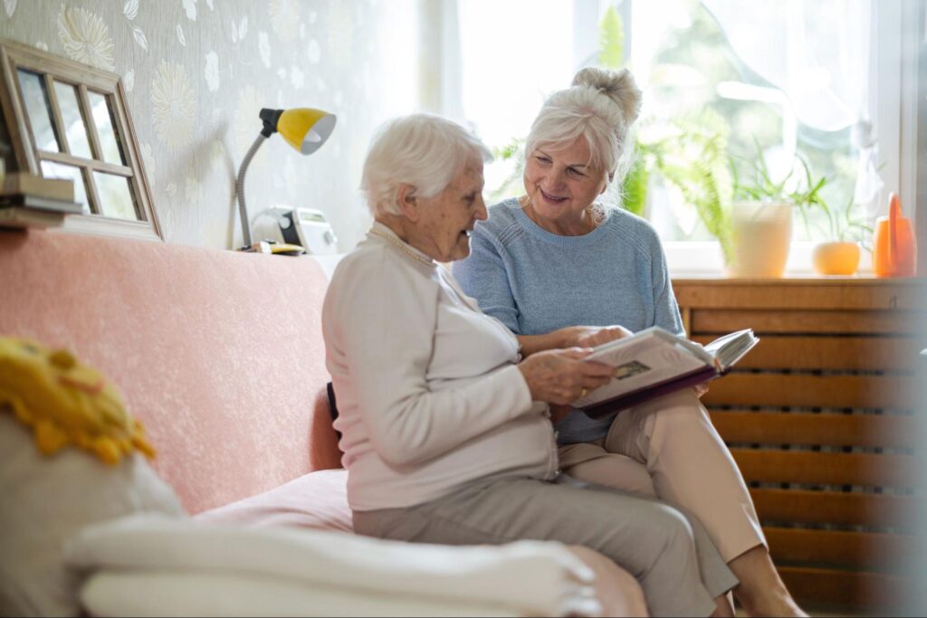 A senior and her adult child looking through a photo album seated on a sofa in a sun-lit room